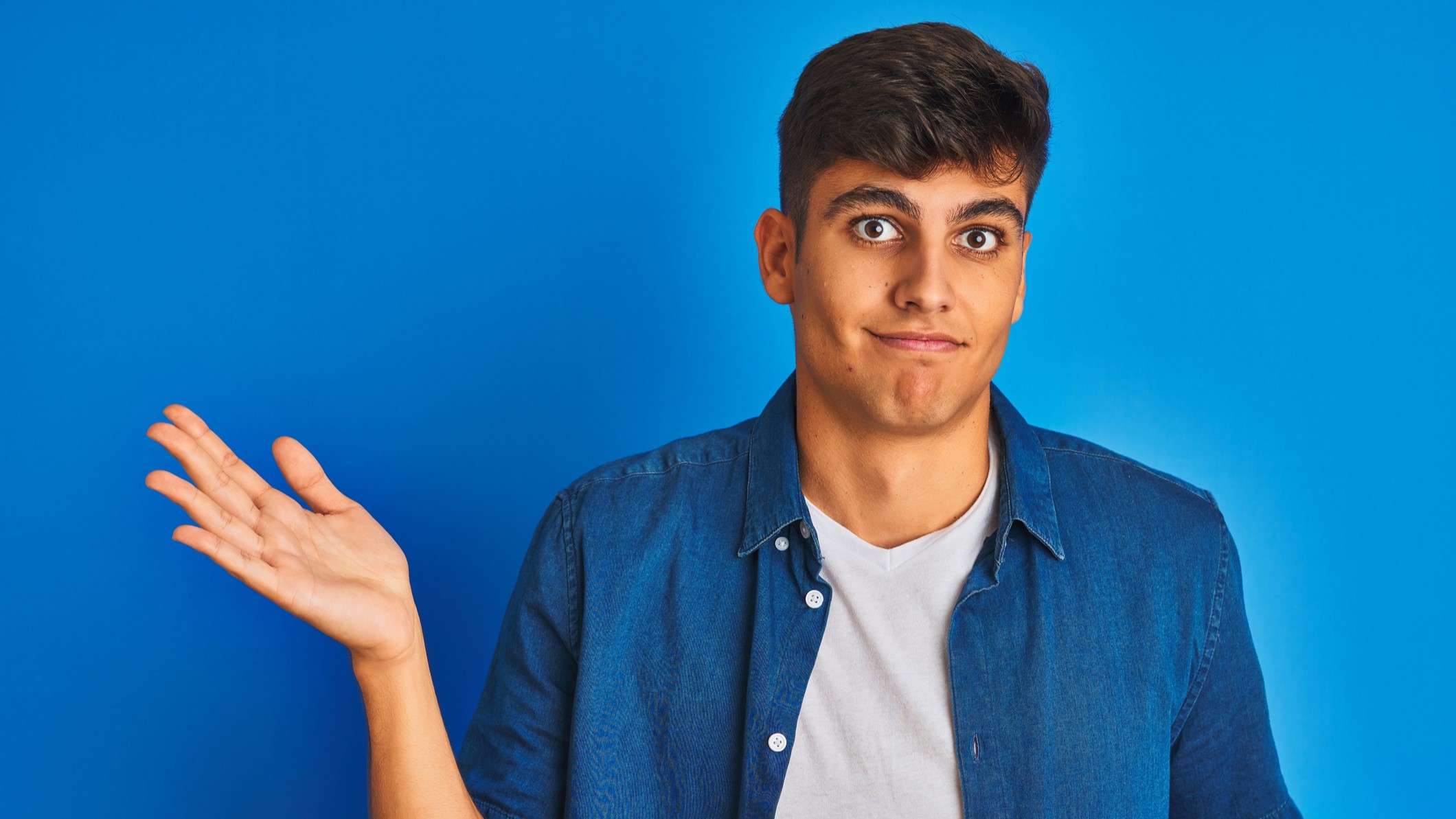 Young man wearing shirt standing over isolated blue background clueless and confused expression with arms and hands raised